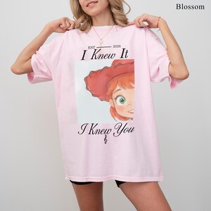 May include: A light pink short-sleeve t-shirt featuring a graphic of a cartoon character with red hair and a red hat. The text reads "EST 2026 I Knew It I Knew You" with a small musical note symbol. The word "Blossom" is printed in the top right corner.