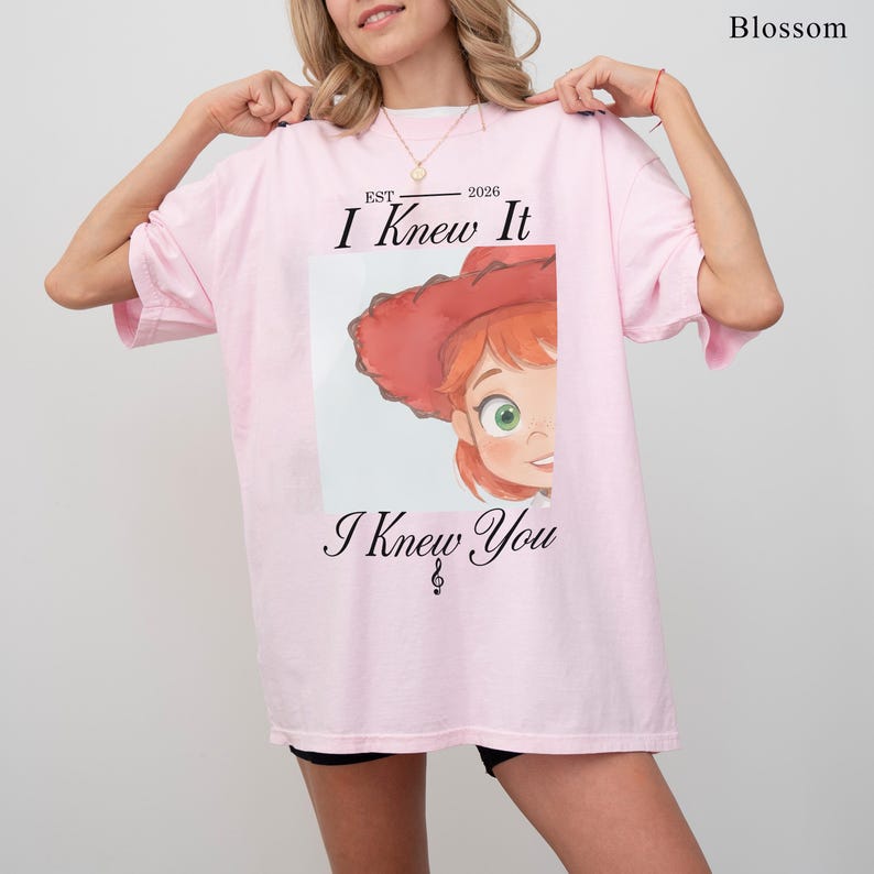 May include: A light pink short-sleeve t-shirt featuring a graphic of a cartoon character with red hair and a red hat. The text reads "EST 2026 I Knew It I Knew You" with a small musical note symbol. The word "Blossom" is printed in the top right corner.