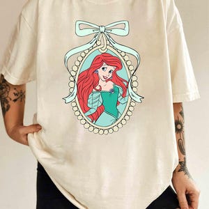 May include: A white t-shirt with a graphic of Ariel, the Little Mermaid, in a pearl-encrusted frame with a blue ribbon bow. The text "Ivory" is visible in the bottom right corner of the image.