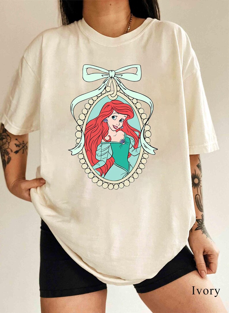 May include: A white t-shirt with a graphic of Ariel, the Little Mermaid, in a pearl-encrusted frame with a blue ribbon bow. The text "Ivory" is visible in the bottom right corner of the image.