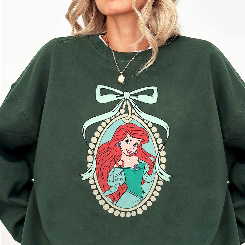 May include: A dark green sweatshirt with a graphic print of Ariel, the Little Mermaid, in a pearl-encrusted frame with a green bow.