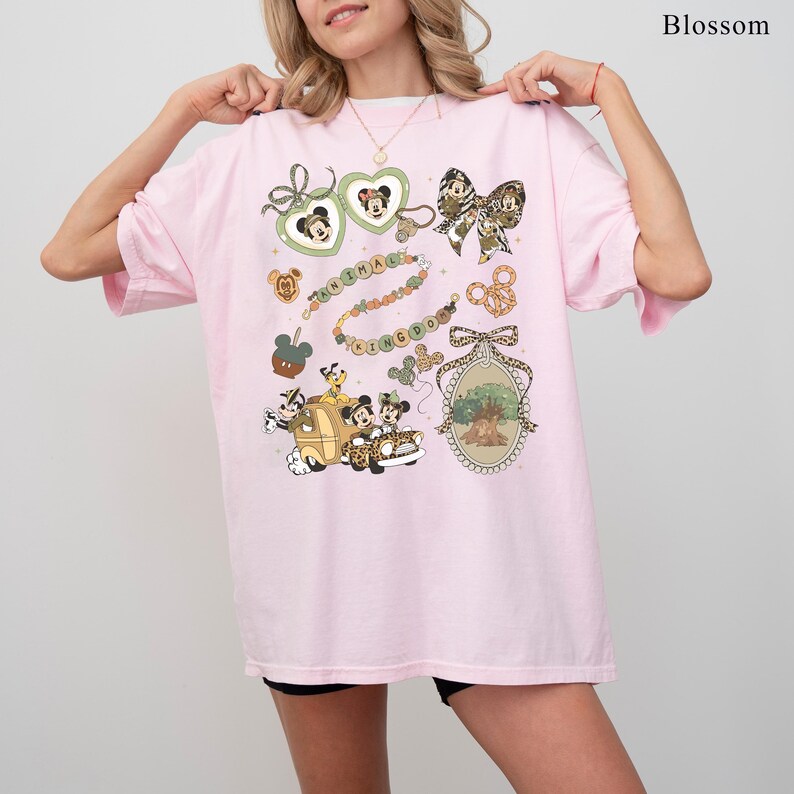 May include: A light pink t-shirt featuring a colorful graphic design. The design includes Mickey Mouse, Minnie Mouse, and other Disney-themed elements. The words "Animal Kingdom" are also visible in the design.