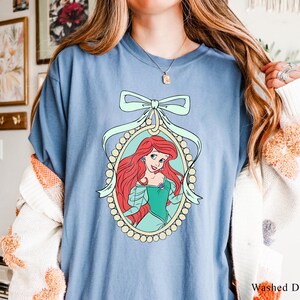 May include: A light blue t-shirt with a graphic of Ariel, the Little Mermaid, in a pearl-encrusted frame with a light blue bow. The graphic is on a light blue background.