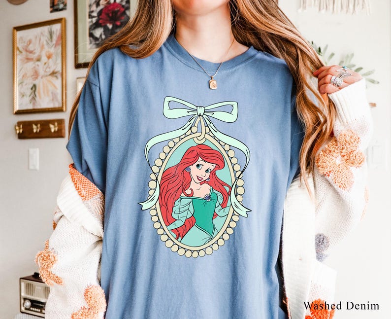 May include: A light blue t-shirt with a graphic of Ariel, the Little Mermaid, in a pearl-encrusted frame with a light blue bow. The graphic is on a light blue background.