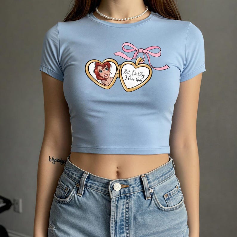 Puede incluir: Una camiseta recortada azul claro con un gr&aacute;fico de un medall&oacute;n en forma de coraz&oacute;n con una imagen de dibujos animados de una mujer con cabello rojo en su interior. El medall&oacute;n tiene el texto "But Dudley I love him".
