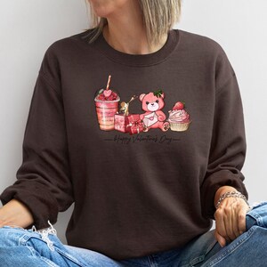 Puede incluir: Una sudadera marr&oacute;n con un oso de peluche rosa, una magdalena rosa, una caja de regalo rosa y un caf&eacute; helado rosa con una pajita en forma de coraz&oacute;n. El texto "Happy Valentine's Day" est&aacute; impreso debajo de la imagen.
