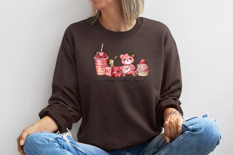 Puede incluir: Una sudadera marr&oacute;n con un oso de peluche rosa, una magdalena rosa, una caja de regalo rosa y un caf&eacute; helado rosa con una pajita en forma de coraz&oacute;n. El texto "Happy Valentine's Day" est&aacute; impreso debajo de la imagen.