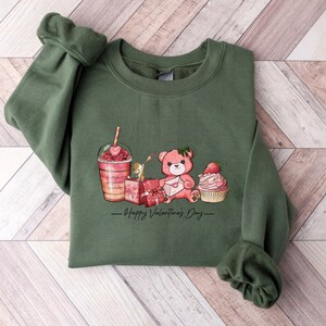 Puede incluir: Sudadera verde con un estampado gr&aacute;fico de un osito de peluche que sostiene una carta, un cupcake con una fresa encima, una caja de regalo y un batido rosa y rojo. El texto "Happy Valentine's Day" est&aacute; impreso debajo del gr&aacute;fico.