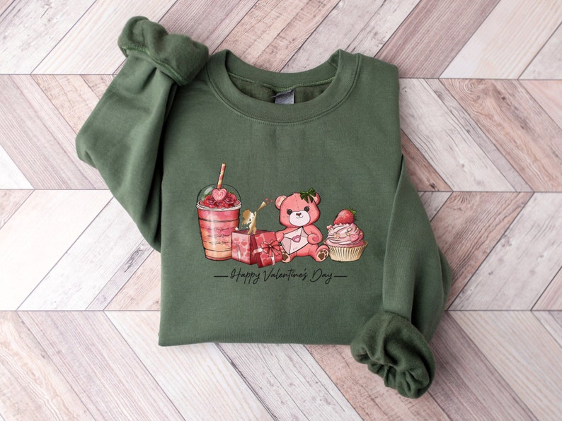 Puede incluir: Sudadera verde con un estampado gr&aacute;fico de un osito de peluche que sostiene una carta, un cupcake con una fresa encima, una caja de regalo y un batido rosa y rojo. El texto "Happy Valentine's Day" est&aacute; impreso debajo del gr&aacute;fico.
