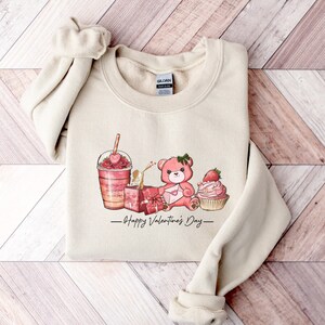 Puede incluir: Una sudadera beige con un dise&ntilde;o gr&aacute;fico de un oso de peluche rosa sosteniendo una carta en forma de coraz&oacute;n, un cupcake rosa con una fresa encima, un batido rosa y naranja con una fresa encima y una caja de regalo rosa. El texto "Happy Valentine's Day" est&aacute; impreso debajo del gr&aacute;fico.