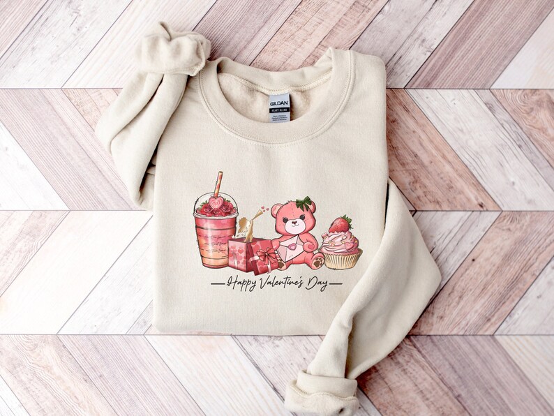 Puede incluir: Una sudadera beige con un dise&ntilde;o gr&aacute;fico de un oso de peluche rosa sosteniendo una carta en forma de coraz&oacute;n, un cupcake rosa con una fresa encima, un batido rosa y naranja con una fresa encima y una caja de regalo rosa. El texto "Happy Valentine's Day" est&aacute; impreso debajo del gr&aacute;fico.