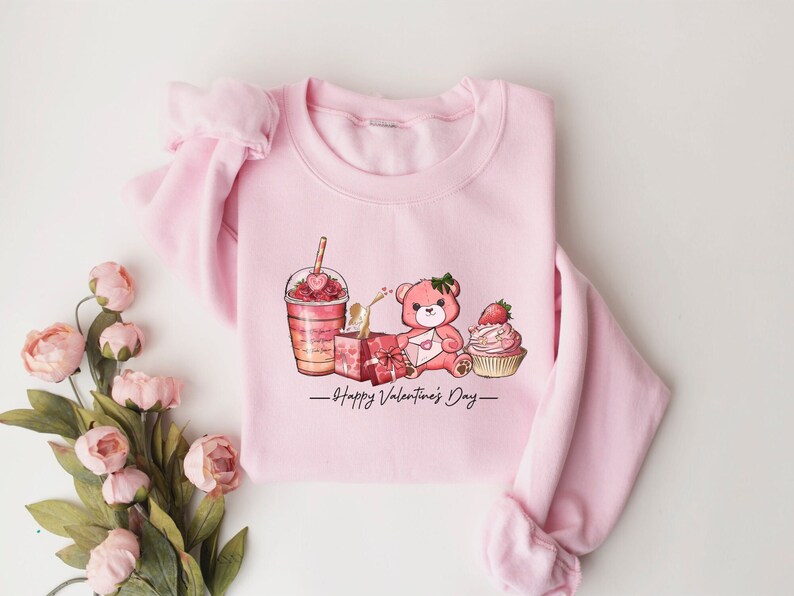 Puede incluir: Sudadera rosa con un dise&ntilde;o gr&aacute;fico de un osito de peluche sosteniendo una carta, un batido, una magdalena y una caja de regalo. El texto "Happy Valentine's Day" est&aacute; impreso debajo del gr&aacute;fico.