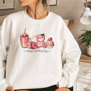 Puede incluir: Sudadera blanca con un oso de peluche rosa, una magdalena rosa, una caja de regalo rosa y un batido rosa con el texto "Happy Valentine's Day" impreso en la parte delantera.