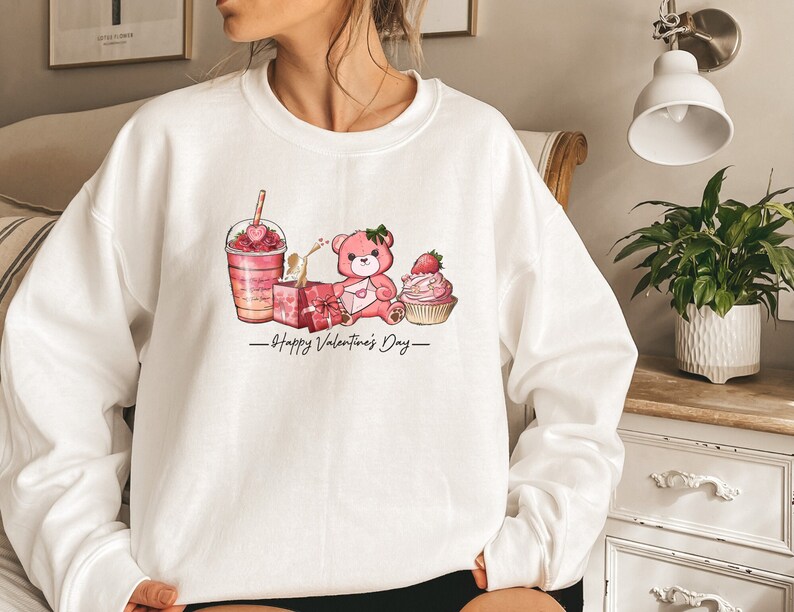 Puede incluir: Sudadera blanca con un oso de peluche rosa, una magdalena rosa, una caja de regalo rosa y un batido rosa con el texto "Happy Valentine's Day" impreso en la parte delantera.
