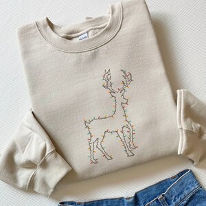 May include: A beige sweatshirt with a brown embroidered reindeer outline decorated with colorful lights. The reindeer is standing on a white background.