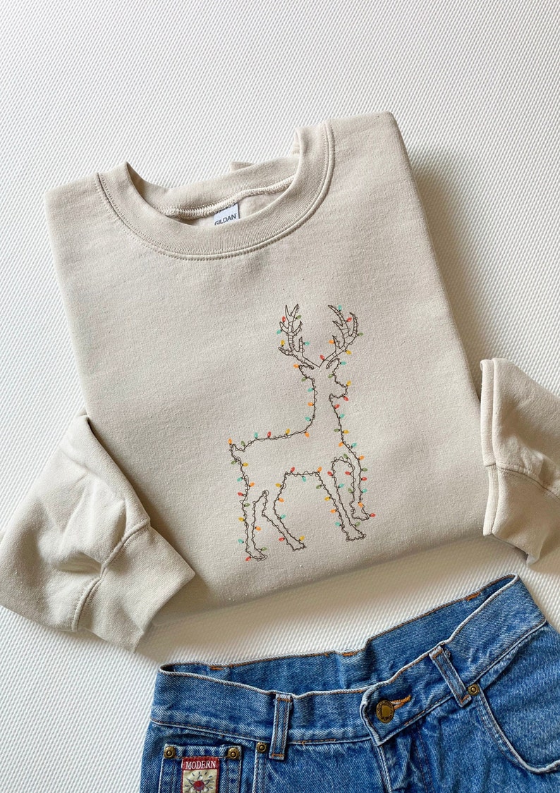 May include: A beige sweatshirt with a brown embroidered reindeer outline decorated with colorful lights. The reindeer is standing on a white background.