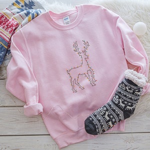 May include: A pink sweatshirt with a white embroidered reindeer design. The reindeer is outlined with colorful lights. The sweatshirt is lying on a white fuzzy surface with a black and white sock with a reindeer pattern.