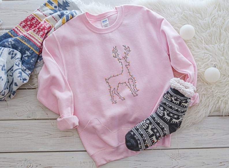 May include: A pink sweatshirt with a white embroidered reindeer design. The reindeer is outlined with colorful lights. The sweatshirt is lying on a white fuzzy surface with a black and white sock with a reindeer pattern.