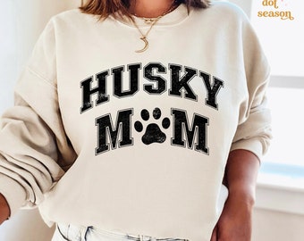 May include: Cream-colored sweatshirt with the text "HUSKY MOM" in a black, distressed, collegiate-style font. A black paw print is centered between the words. The sweatshirt is a casual style.