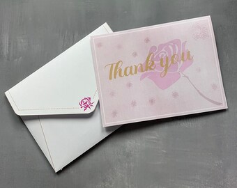 May include: A light pink thank you card featuring a large rose illustration and gold script text, paired with a white envelope that has a small pink rose icon on the flap. The card has a thin border and scattered small rose motifs across the surface.