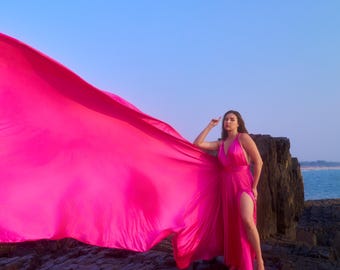 May include: A bright fuchsia dress with a flowing train is shown. The dress has a high slit and a halter neckline. The model is standing on a rocky outcrop near the sea, with a bright blue sky in the background.