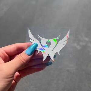 Puede incluir: Una pegatina de vinilo blanco con un dise&ntilde;o de coraz&oacute;n y alas. El coraz&oacute;n est&aacute; relleno de colores arco&iacute;ris iridiscentes.