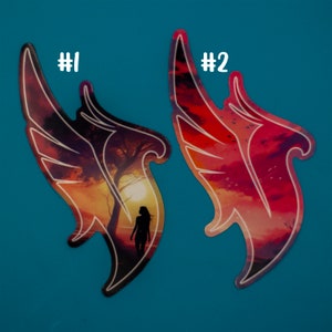 May include: Two colorful stickers with a wing design. The stickers feature a sunset scene with a silhouette of a person standing in front of a tree. The stickers are labeled #1 and #2.