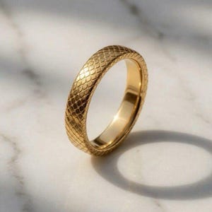 Può includere: Un anello a fascia in oro caratterizzato da una superficie testurizzata con un motivo a scaglie ripetuto. L'anello presenta un interno lucido e una finitura metallica, che riflette la luce sulla superficie esterna per evidenziare le piccole scaglie a rombo.