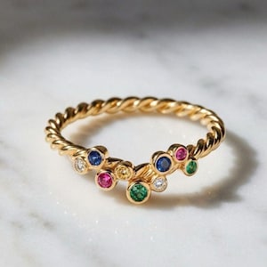 May include: A gold ring with a twisted band featuring a cluster of small, round, multi-coloured gemstones including blue, pink, green, yellow, and clear stones set in a staggered arrangement along the front of the band.