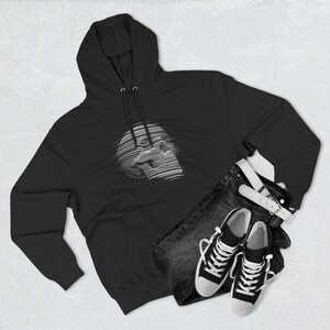 May include: Black hoodie featuring a white, linear skull design. The hoodie is styled with dark denim jeans, a white belt, and black and white trainers. The ensemble is arranged on a light grey surface.