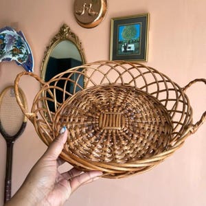 May include: A round, light brown woven wicker basket featuring a decorative lattice-style openwork rim and two integrated side handles. The base of the basket is constructed with a tight, circular weave pattern, providing a sturdy foundation.