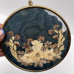 May include: A circular wall hanging featuring a brass frame and glass front, enclosing a dark teal fabric background decorated with an arrangement of pressed, dried cream and tan flowers and grasses, topped with a small brass hanging loop.