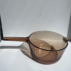 May include: A vintage amber-colored glass saucepan with a long handle. The saucepan has a rounded bowl shape and a pouring spout. The handle is attached to the side of the pan. The glass is transparent, allowing the contents to be seen.