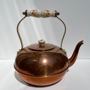 May include: A polished copper tea kettle with a rounded body and a curved spout. The handle features a white ceramic grip decorated with small orange berries and green leaves. A matching white ceramic knob sits atop the removable lid.