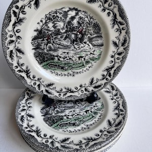 May include: A stack of white ceramic plates featuring a black and white hunting scene. Each plate has a decorative black leaf and vine border. The plates are stacked, showcasing the design.