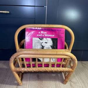 May include: A vintage rattan magazine rack holding a vinyl record album. The album cover is pink and features the text "A Man and a Woman". The rack is made of light brown woven rattan.