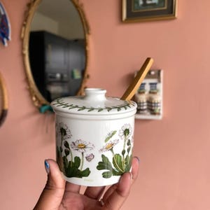May include: A white ceramic lidded jar featuring a botanical illustration of daisy flowers with green leaves and small butterflies. The lid has a green leaf border pattern and a central knob. A wooden spoon is inserted into the side of the lid.