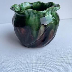 May include: A small ceramic planter with a ruffled rim. The exterior has a glossy glaze in shades of green and brown, creating a marbled effect. The rounded shape and unique colour palette make it a decorative piece for plants.