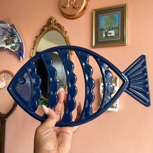 May include: A dark blue, fish-shaped trivet with a glossy finish. The design features a skeletal structure with wavy, vertical cutouts representing the body segments, a defined head with a circular eye, and a fanned tail section.