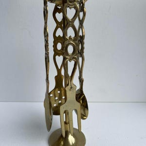 May include: A brass fireplace tool set with a stand. The stand has a round base and a decorative openwork design with heart shapes. The set includes a poker, shovel, and other tools. The tools are made of brass and have a shiny finish.