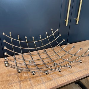May include: A chrome-finished metal fruit basket with a curved, grid-like design. The basket features small, spherical accents along the edges and is displayed on a light-colored wooden surface. The basket is a decorative item.