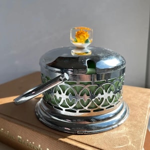 May include: A vintage silver-plated sugar bowl with a handle and a decorative openwork design. The bowl features a green interior and is topped with a clear, square-shaped lid containing a yellow flower. The bowl is resting on a brown book.