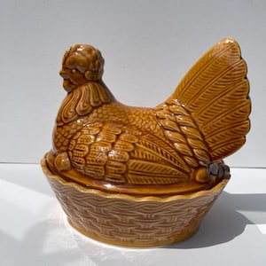 May include: A glossy, honey-brown ceramic hen-shaped lidded dish. The lid is moulded in the shape of a sitting hen with detailed feather textures, while the base is designed to resemble a woven basket with a scalloped rim edge.