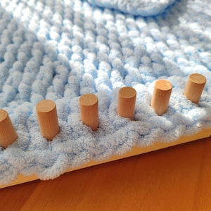 Può includere: Primo piano di una coperta lavorata a maglia grossa azzurra in lavorazione su un telaio di legno. Il telaio ha pioli di legno distanziati uniformemente. La coperta è realizzata con filato morbido e soffice.