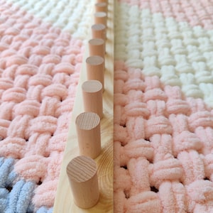 Op de afbeelding: Een lange, rechthoekige plank van lichtgekleurd hout met een enkele rij gelijkmatig verdeelde, cilindrische houten pinnen die omhoog steken. De plank ligt op een zachte, grofgebreide deken met een geweven patroon in pastelroze, wit en blauw.