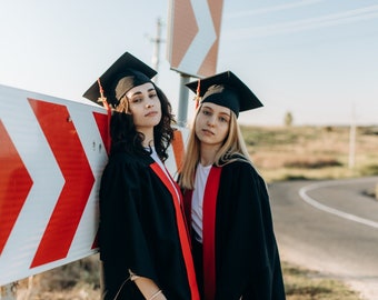 Puede incluir: Dos graduadas con birretes y togas negras con detalles rojos posan cerca de una señal de tráfico con chevrones rojos y blancos. Las graduadas están de pie junto a una carretera con un fondo borroso.