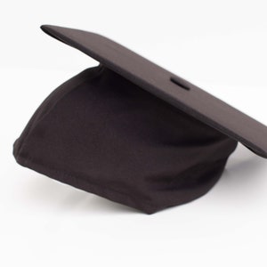 Może przedstawiać: Czarny czapkę doktorską z kwadratowym wierzchołkiem i czarną frędzlą. Czapka jest wykonana z tkaniny i leży na białym tle.