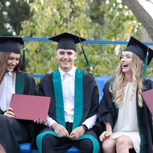 Op de afbeelding: Drie afgestudeerden in zwarte mutsen en toga's, zittend op een blauwe bank, met diploma's in de hand. De afgestudeerden lachen en glimlachen.