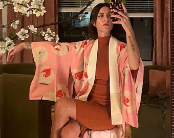 May include: A pink kimono-style robe with a white collar and crane design. The robe is worn over a rust-colored dress. Black flats with an eye design are visible. The background includes a mirror, green sofa, and floral arrangement.
