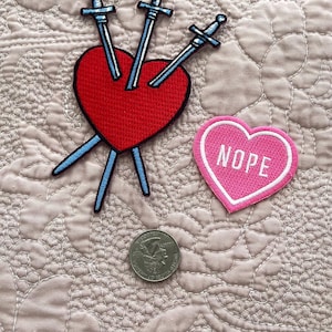 Könnte beinhalten: Zwei gestickte Bügelbilder. Eines ist ein rotes Herz, das von drei Schwertern durchbohrt wird. Das andere ist ein pinkfarbenes Herz mit dem Wort "NOPE" in weißen Buchstaben.